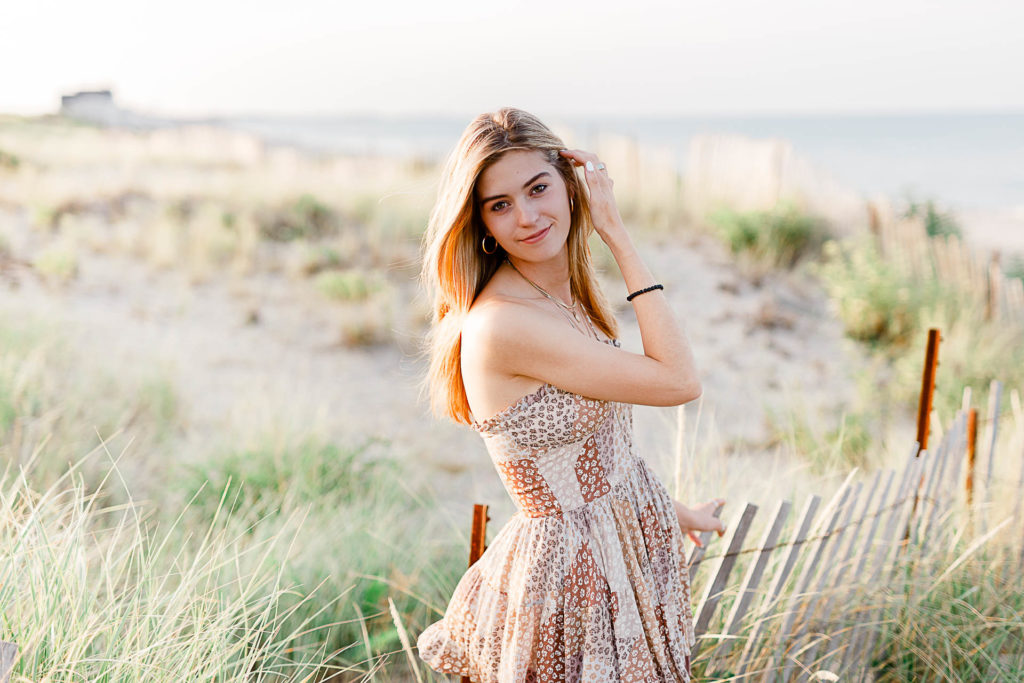 Photo by Cohasset senior portrait photographer Christina Runnals | High school senior girl standing in front of beach grass and a fence with water in the background