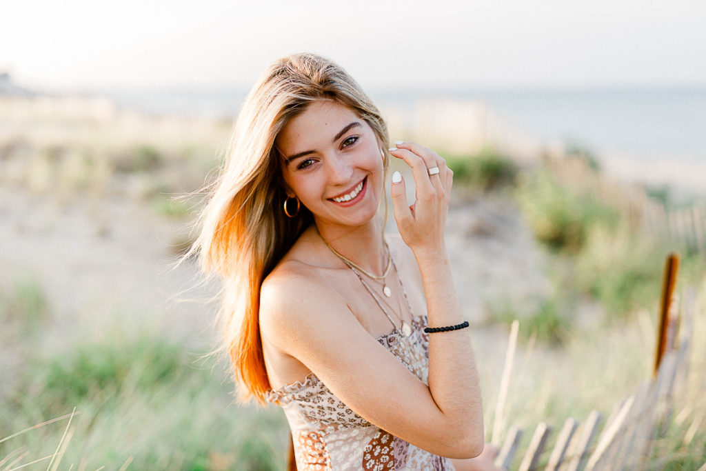 Photo by Cohasset senior portrait photographer Christina Runnals | High school senior girl standing in front of beach grass and a fence with water in the background