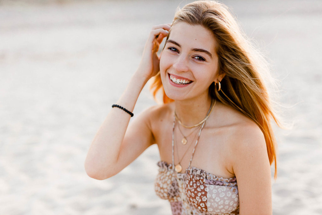 Photo by Cohasset senior portrait photographer Christina Runnals | High school senior girl sitting and smiling on the beach at sunset 