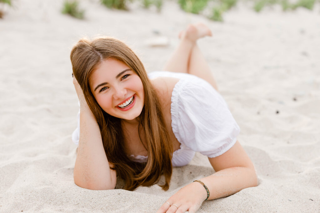 Girl in white dress laying on stomach on the beach smiling with her hand in her hair