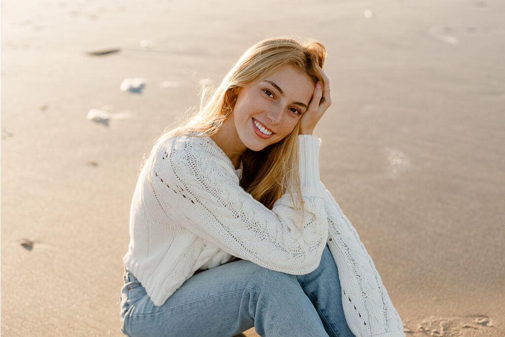 Fall senior pictures in Massachusetts featuring Stella in cream colored chunky knit sweater and blue jeans sitting on the beach.