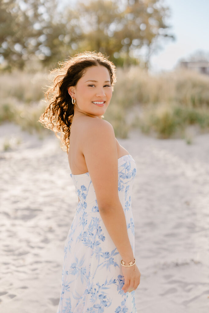 Lexington MA senior pictures taken on the South Shore at a beach.  Featuring a senior with long curly dark brown hair in a long white dress with a blue floral pattern.