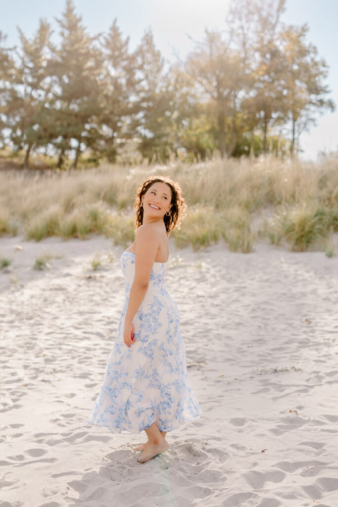 Lexington MA senior pictures taken on the South Shore at a beach.  Featuring a senior with long curly dark brown hair in a long white dress with a blue floral pattern.