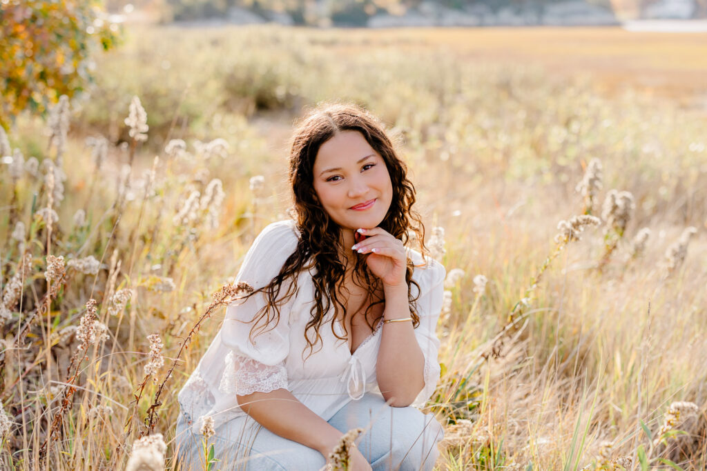 Lexington MA senior pictures taken on the South Shore at a beach.  Featuring a senior with long curly dark brown hair in jeans and a white blouse with lace detail.  Surrounded by autumn foliage and a gorgeous field.