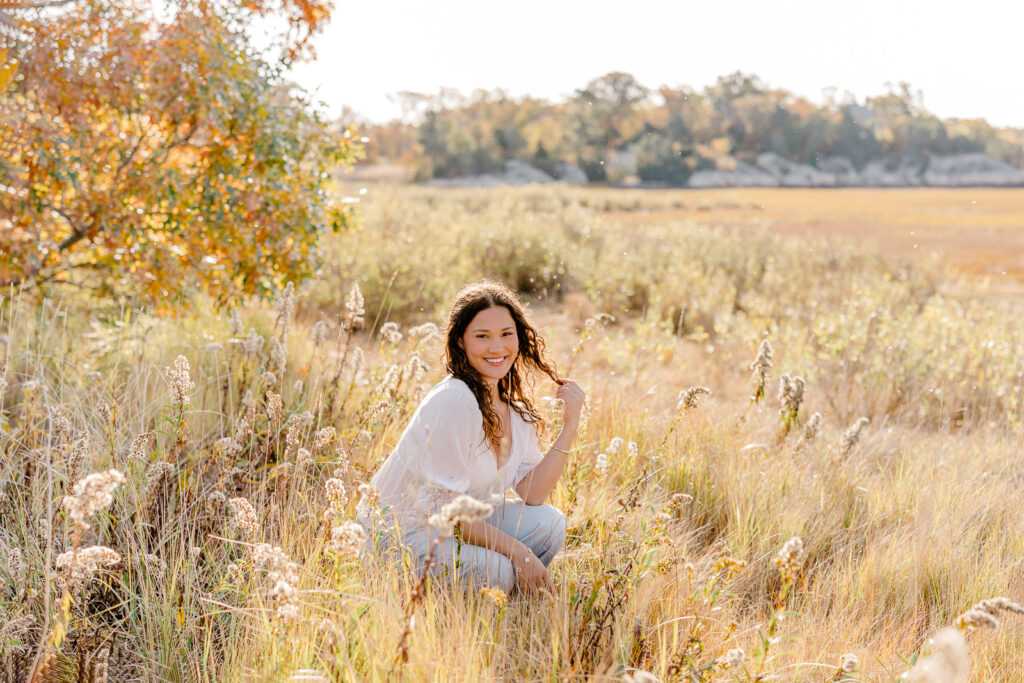 Lexington MA senior pictures taken on the South Shore at a beach.  Featuring a senior with long curly dark brown hair in jeans and a white blouse with lace detail.  Surrounded by autumn foliage and a gorgeous field.
