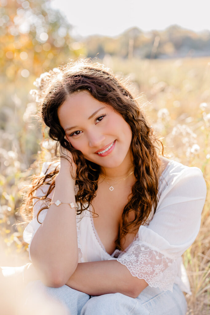 Lexington MA senior pictures taken on the South Shore at a beach.  Featuring a senior with long curly dark brown hair in jeans and a white blouse with lace detail.  Candid, natural posing.  Surrounded by autumn foliage and a gorgeous field.