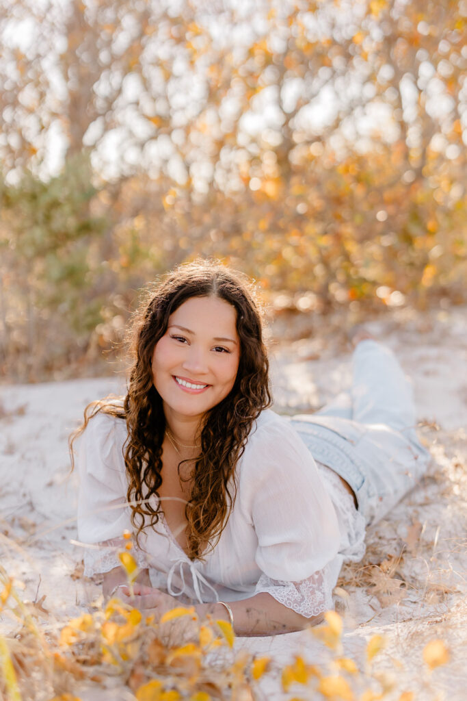 Lexington MA senior pictures taken on the South Shore at a beach.  Featuring a senior with long curly dark brown hair in jeans and a white blouse with lace detail.  Candid, natural posing.  Surrounded by autumn foliage and a gorgeous field.