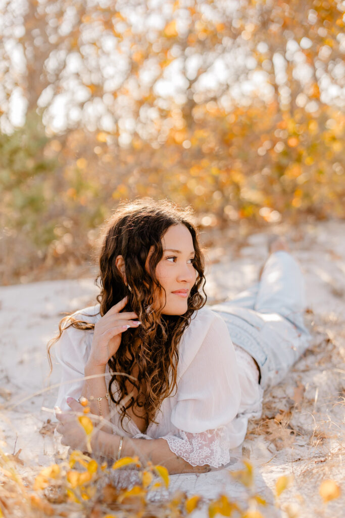 Lexington MA senior pictures taken on the South Shore at a beach.  Featuring a senior with long curly dark brown hair in jeans and a white blouse with lace detail.  Candid, natural posing.  Surrounded by autumn foliage and a gorgeous field.