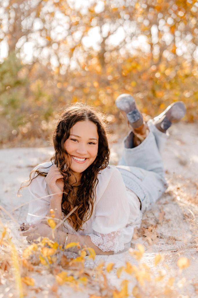 Lexington MA senior pictures taken on the South Shore at a beach.  Featuring a senior with long curly dark brown hair in jeans and a white blouse with lace detail and brown cowboy boots.  Candid, natural posing.  Surrounded by autumn foliage and a gorgeous field.